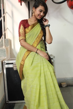 A woman is standing indoors wearing a traditional green and yellow sari with a red blouse, accessorized with earrings, a watch, and a bindi. Her hair is styled in a long braid, and she is smiling gently while posing with one hand touching her braid. The background includes a staircase and a black equipment case.