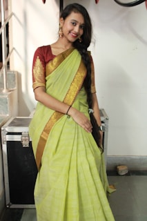 A woman in a traditional outfit stands indoors. She is wearing a lime green sari with gold embroidery and a maroon blouse. Her hair is styled in a long braid down her shoulder, and she accessorizes with gold earrings. The setting includes a metallic case and a staircase in the background.