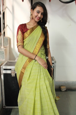A woman in a traditional outfit stands indoors. She is wearing a lime green sari with gold embroidery and a maroon blouse. Her hair is styled in a long braid down her shoulder, and she accessorizes with gold earrings. The setting includes a metallic case and a staircase in the background.
