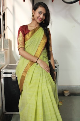 A woman in a traditional outfit stands indoors. She is wearing a lime green sari with gold embroidery and a maroon blouse. Her hair is styled in a long braid down her shoulder, and she accessorizes with gold earrings. The setting includes a metallic case and a staircase in the background.