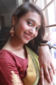 A young woman is smiling while leaning against a window with bars. She is wearing a traditional Indian outfit consisting of a maroon blouse and a green and gold sari. Her accessories include large, ornate earrings and several rings. Her makeup is subtle with an emphasis on her eyes and lips.