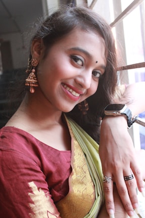 A young woman is smiling while leaning against a window with bars. She is wearing a traditional Indian outfit consisting of a maroon blouse and a green and gold sari. Her accessories include large, ornate earrings and several rings. Her makeup is subtle with an emphasis on her eyes and lips.