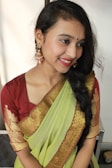 A person is wearing a traditional Indian saree that is predominantly green with gold patterns along the border and paired with a maroon blouse. The person has braided hair and is adorned with large, intricate earrings. The background is simple and light, highlighting the subject.