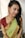 A person is wearing a traditional Indian saree that is predominantly green with gold patterns along the border and paired with a maroon blouse. The person has braided hair and is adorned with large, intricate earrings. The background is simple and light, highlighting the subject.