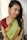 A person is wearing a traditional Indian saree that is predominantly green with gold patterns along the border and paired with a maroon blouse. The person has braided hair and is adorned with large, intricate earrings. The background is simple and light, highlighting the subject.