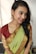 A person is wearing a traditional Indian saree that is predominantly green with gold patterns along the border and paired with a maroon blouse. The person has braided hair and is adorned with large, intricate earrings. The background is simple and light, highlighting the subject.