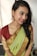 A person is wearing a traditional Indian saree that is predominantly green with gold patterns along the border and paired with a maroon blouse. The person has braided hair and is adorned with large, intricate earrings. The background is simple and light, highlighting the subject.
