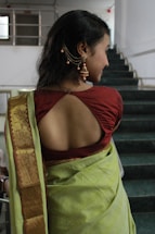 A woman is seen wearing a traditional outfit, consisting of a red blouse with an open back design and a green sari with a gold border. Her hair is styled with elegant jewelry, including earrings and a decorative hair accessory. She stands on a staircase in a well-lit indoor setting.