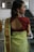 A woman wearing traditional attire stands with her back facing the camera. She is wearing a green sari with a patterned gold border and a maroon blouse featuring an open back design. Her hair is styled to one side, adorned with gold earrings featuring intricate designs.