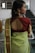 A woman wearing traditional attire stands with her back facing the camera. She is wearing a green sari with a patterned gold border and a maroon blouse featuring an open back design. Her hair is styled to one side, adorned with gold earrings featuring intricate designs.
