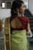 A woman wearing traditional attire stands with her back facing the camera. She is wearing a green sari with a patterned gold border and a maroon blouse featuring an open back design. Her hair is styled to one side, adorned with gold earrings featuring intricate designs.