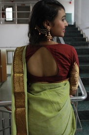A woman wearing traditional attire stands with her back facing the camera. She is wearing a green sari with a patterned gold border and a maroon blouse featuring an open back design. Her hair is styled to one side, adorned with gold earrings featuring intricate designs.