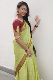 A woman is dressed in a traditional Indian sari, featuring a light green color with a golden border. She is standing against a plain white wall, smiling, and is adorned with gold jewelry, including earrings, a black wristwatch, and a bindi on her forehead. Her long black hair is styled with a braid over one shoulder.