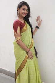 A woman is dressed in a traditional Indian sari, featuring a light green color with a golden border. She is standing against a plain white wall, smiling, and is adorned with gold jewelry, including earrings, a black wristwatch, and a bindi on her forehead. Her long black hair is styled with a braid over one shoulder.
