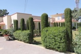 A modern villa with a cream exterior and red roofing is surrounded by neatly trimmed hedges and tall, cylindrical shrubs. The driveway is paved with patterned stones, and a large potted plant adds a decorative touch near the entrance. The sky is clear and bright, suggesting a sunny day.