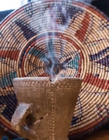 A rustic, textured incense burner emitting smoke is placed in front of a colorful, woven basket with intricate geometric patterns. The composition highlights the earthy tones and cultural craftsmanship.