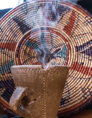 A rustic, textured incense burner emitting smoke is placed in front of a colorful, woven basket with intricate geometric patterns. The composition highlights the earthy tones and cultural craftsmanship.