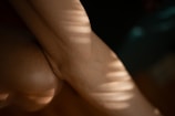 Soft sunlight creates patterned shadows on a close-up of skin, highlighting the texture with alternating light and shadow, conveying a sense of warmth and intimacy.