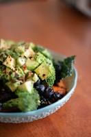 A bowl filled with a variety of fresh, healthy ingredients including chopped avocado, broccoli, blueberries, and what appears to be kale. The dish is garnished with red pepper flakes and sits on a wooden surface.