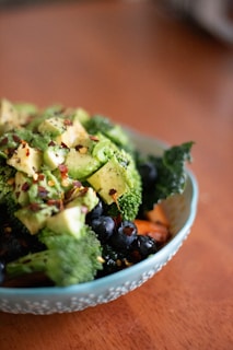 A bowl filled with a variety of fresh, healthy ingredients including chopped avocado, broccoli, blueberries, and what appears to be kale. The dish is garnished with red pepper flakes and sits on a wooden surface.