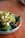 A bowl filled with a variety of fresh, healthy ingredients including chopped avocado, broccoli, blueberries, and what appears to be kale. The dish is garnished with red pepper flakes and sits on a wooden surface.