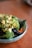 A bowl filled with a variety of fresh, healthy ingredients including chopped avocado, broccoli, blueberries, and what appears to be kale. The dish is garnished with red pepper flakes and sits on a wooden surface.