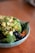 A bowl filled with a variety of fresh, healthy ingredients including chopped avocado, broccoli, blueberries, and what appears to be kale. The dish is garnished with red pepper flakes and sits on a wooden surface.
