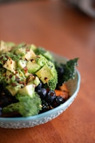 A bowl filled with a variety of fresh, healthy ingredients including chopped avocado, broccoli, blueberries, and what appears to be kale. The dish is garnished with red pepper flakes and sits on a wooden surface.