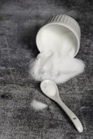 A white ceramic ramekin lies on its side, spilling a mound of fine white granulated sugar onto a textured dark gray surface. A white ceramic spoon is nearby, also dusted with sugar.
