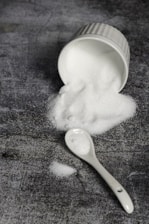 A white ceramic ramekin lies on its side, spilling a mound of fine white granulated sugar onto a textured dark gray surface. A white ceramic spoon is nearby, also dusted with sugar.