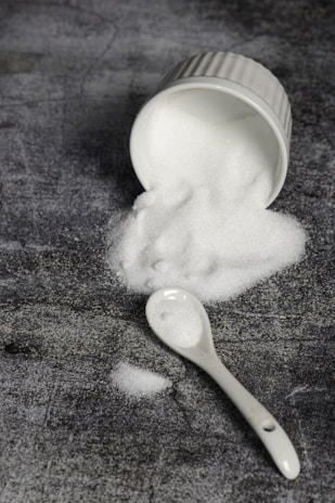 A white ceramic ramekin lies on its side, spilling a mound of fine white granulated sugar onto a textured dark gray surface. A white ceramic spoon is nearby, also dusted with sugar.