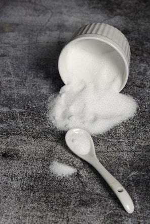 A white ceramic ramekin lies on its side, spilling a mound of fine white granulated sugar onto a textured dark gray surface. A white ceramic spoon is nearby, also dusted with sugar.