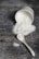 A white ceramic ramekin lies on its side, spilling a mound of fine white granulated sugar onto a textured dark gray surface. A white ceramic spoon is nearby, also dusted with sugar.