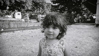 A young child with curly hair stands outdoors near railway tracks. The background features trees, a pathway, and a house.