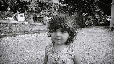 A young child with curly hair stands outdoors near railway tracks. The background features trees, a pathway, and a house.