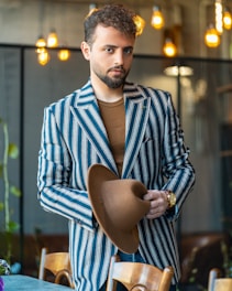 A man with a beard is holding a brown hat in his hands, dressed in a stylish, striped blue and white blazer over a brown shirt. He is standing in a well-lit room with modern decor and warm, ambient lighting hanging from the ceiling.