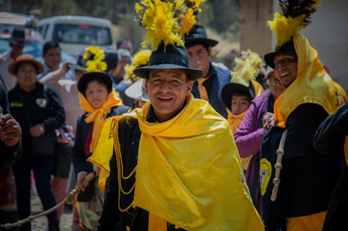 A group of people is dressed in vibrant traditional attire, prominently featuring bright yellow shawls and black hats adorned with yellow feathers. They are gathered closely together, creating a festive and lively atmosphere. The background includes a few vehicles and more people, suggesting a public or community celebration.