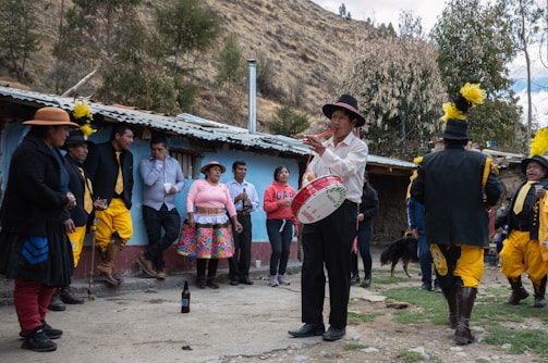 A group of people gathers outdoors near a rural building. One man plays a drum while the rest are dressed in colorful traditional attire, with some wearing hats. The setting appears to be festive, possibly a cultural celebration or event in a mountainous area with trees and a rugged landscape.