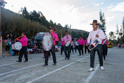 A group of musicians wearing pink shirts and black pants with wide-brimmed hats are playing drums and marching in an outdoor setting. A crowd of people can be seen seated on colorful bleachers in the background, and the environment is surrounded by trees and hills under a partly cloudy sky.