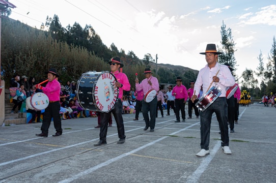 A group of musicians wearing pink shirts and black pants with wide-brimmed hats are playing drums and marching in an outdoor setting. A crowd of people can be seen seated on colorful bleachers in the background, and the environment is surrounded by trees and hills under a partly cloudy sky.