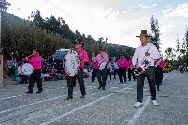 A group of musicians wearing pink shirts and black pants with wide-brimmed hats are playing drums and marching in an outdoor setting. A crowd of people can be seen seated on colorful bleachers in the background, and the environment is surrounded by trees and hills under a partly cloudy sky.