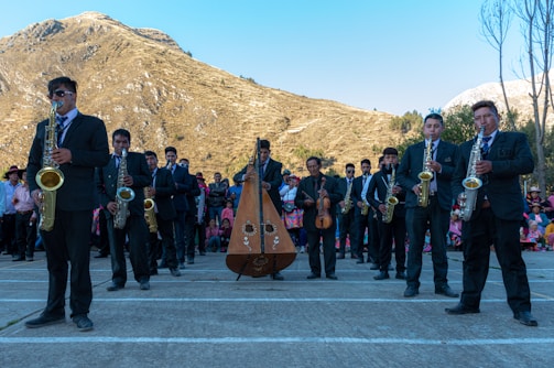 A group of musicians wearing formal attire performs outdoors in front of a scenic mountain landscape. The ensemble includes saxophonists and a harp player, while a crowd observes in the background, some dressed in colorful traditional clothing. The setting is bright with clear skies.