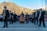 A group of musicians wearing formal attire performs outdoors in front of a scenic mountain landscape. The ensemble includes saxophonists and a harp player, while a crowd observes in the background, some dressed in colorful traditional clothing. The setting is bright with clear skies.