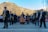 A group of musicians wearing formal attire performs outdoors in front of a scenic mountain landscape. The ensemble includes saxophonists and a harp player, while a crowd observes in the background, some dressed in colorful traditional clothing. The setting is bright with clear skies.