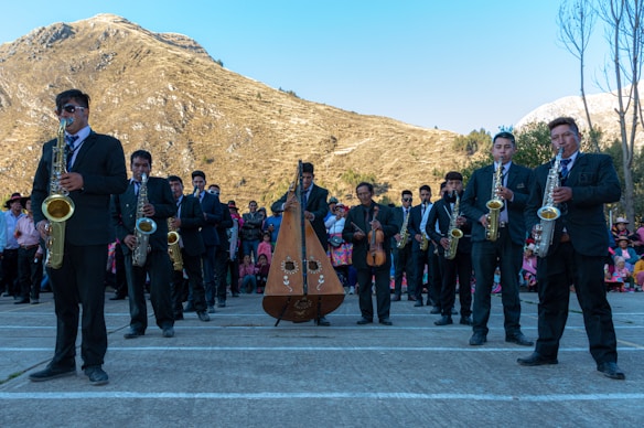 A group of musicians wearing formal attire performs outdoors in front of a scenic mountain landscape. The ensemble includes saxophonists and a harp player, while a crowd observes in the background, some dressed in colorful traditional clothing. The setting is bright with clear skies.