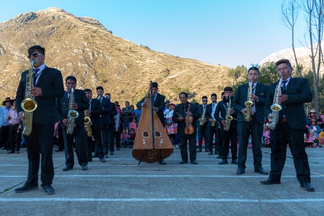 A group of musicians wearing formal attire performs outdoors in front of a scenic mountain landscape. The ensemble includes saxophonists and a harp player, while a crowd observes in the background, some dressed in colorful traditional clothing. The setting is bright with clear skies.