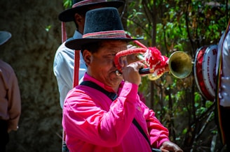 A man wearing a bright pink shirt and a traditional black hat with red accents plays a brass trumpet adorned with red and white tassels. Another person is partially visible in the background, along with a drum, amidst greenery.