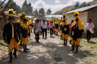 A group of people dressed in vibrant traditional attire with yellow accents and tall black hats with yellow plumes are standing in an outdoor setting. They are gathered along a dirt path with houses on either side. Some are holding drums or other musical instruments. Trees and hills are visible in the background under a partly cloudy sky.
