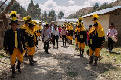 A group of people dressed in vibrant traditional attire with yellow accents and tall black hats with yellow plumes are standing in an outdoor setting. They are gathered along a dirt path with houses on either side. Some are holding drums or other musical instruments. Trees and hills are visible in the background under a partly cloudy sky.