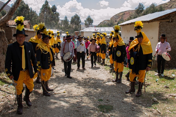 A group of people dressed in vibrant traditional attire with yellow accents and tall black hats with yellow plumes are standing in an outdoor setting. They are gathered along a dirt path with houses on either side. Some are holding drums or other musical instruments. Trees and hills are visible in the background under a partly cloudy sky.